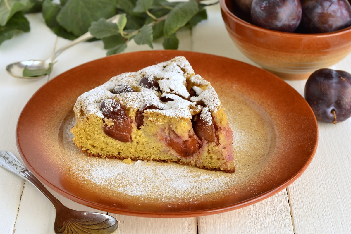 Porție de prăjitură de post cu prune pe o farfurie maro, alături de un bol cu prune