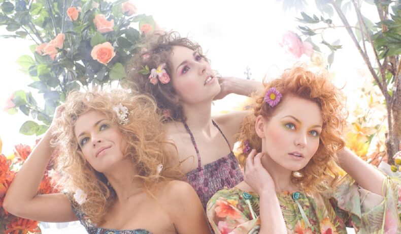 Trei prietene care se află lângă o tufă de trandafiri, au rochii colorate, iar în păr și-au prins diferite flori