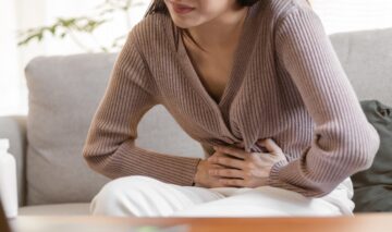 O femeie care stă pe canapea și își ține mâinile pe burtă pentru a ilustra cele 3 remedii care rezolvă constipația într-o singură zi
