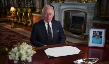 Regele Charles în timp ce stă la un birou alături de portretul Reginei Elisabeta a II-a