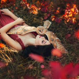O femeie frumoasă care poartă o rochie roșie și stă întinsă pe un covor de frunze alături de o bufniță care o îmbrățișează pentru a ilustra care sunt cele mai empatice zodii