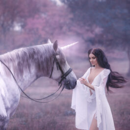 O femeie frumoasă, brunetă, îmbrăcată într-o rochie albă, vaporoasă, care atinge un unicorn