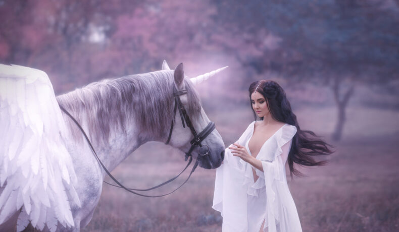 O femeie frumoasă, brunetă, îmbrăcată într-o rochie albă, vaporoasă, care atinge un unicorn