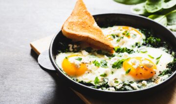 O farfurie plină cu două ouă ochiuri cu brânză feta și servite cu o felie de pâine