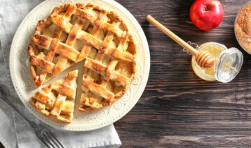 Tartă cu mere porționată pe platou, alături de un borcan cu miere și lingură de lemn, un măr și un bol cu zahăr brun