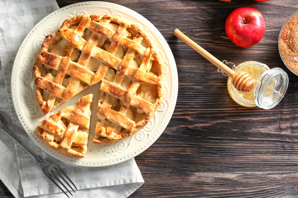 Tartă cu mere porționată pe platou, alături de un borcan cu miere și lingură de lemn, un măr și un bol cu zahăr brun