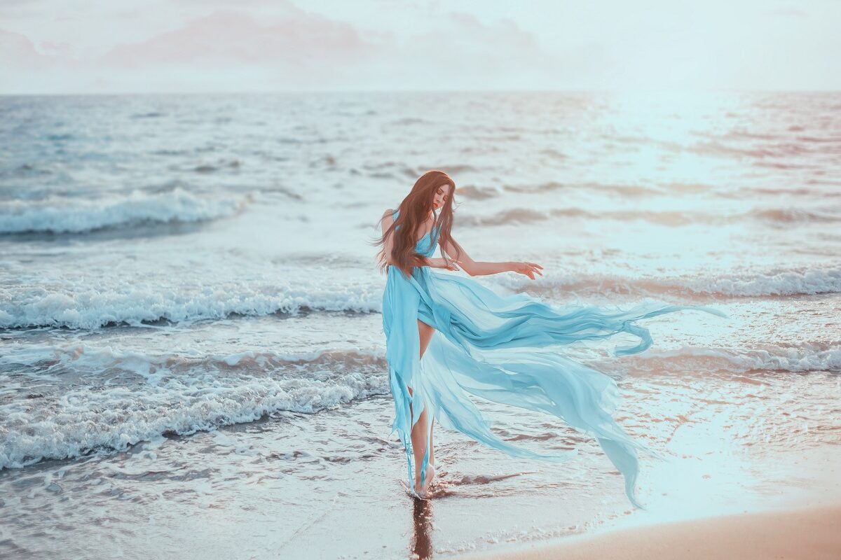 O femeie frumoasă care poartă o rochie albastră și pozează pe malul mării pentru a ilustra una dintre cele trei zodii care au parte de ghinion în ziua de 24 septembrie 2023