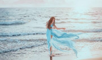 O femeie frumoasă care poartă o rochie albastră și pozează pe malul mării pentru a ilustra una dintre cele trei zodii care au parte de ghinion în ziua de 24 septembrie 2023