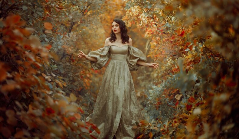 Fată frumoasă îmbrăcată într-o rochie lungă stă într-o pădure plină de frunze