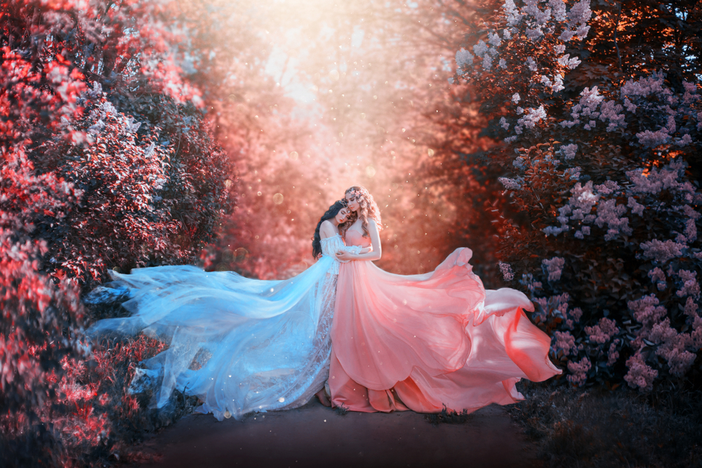 Două fete cu rochii lungi de voal stau îmbrățișate într-o pădure plină de flori colorate