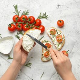 Două mâini de femeie care întind brânză pe pâine cu cuțitul, alături sunt mai multe roșii cherry