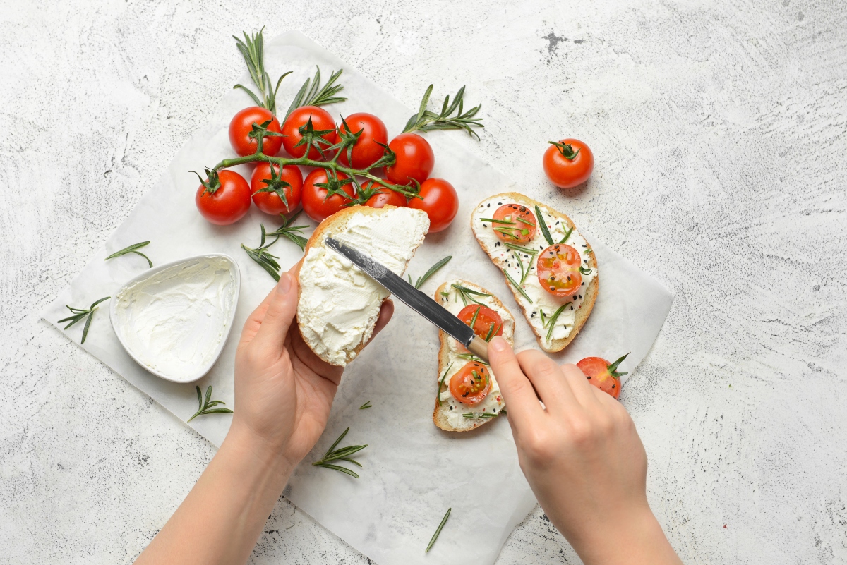 Două mâini de femeie care întind brânză pe pâine cu cuțitul, alături sunt mai multe roșii cherry