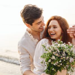 Cuplu fericit pe plajă, cei doi se țin în brațe și ea ține în mână un buchet de flori
