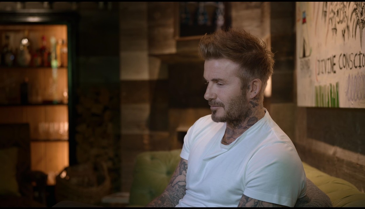 David Beckham, într-un tricou alb, pe o canapea, cu capul plecat