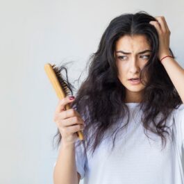 O femeie privește îngrozită o perie de păr plină de fire rupte în timp ce așază o palmă peste o latură a părului