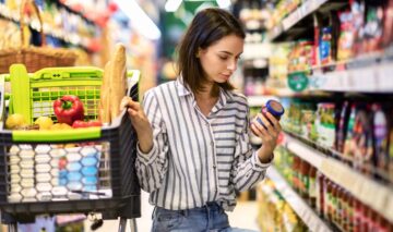 O femeie fruomoasă care citește capcana de pe eticheta alimentelor și se sprijină de un cărucior de cumpărături
