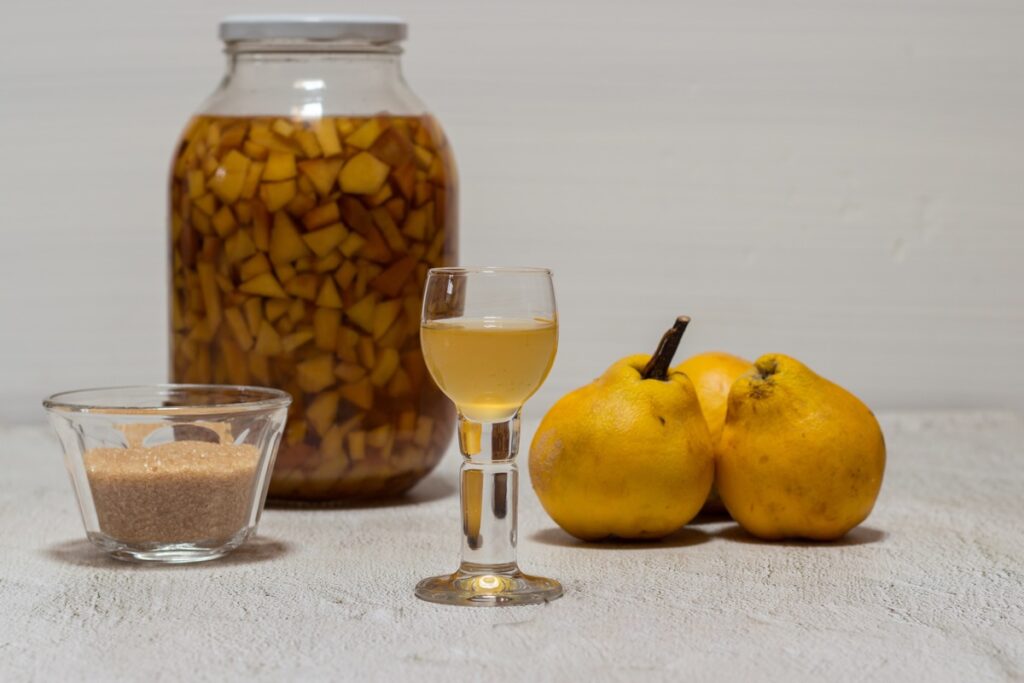 Cuburi de gutui la macert în borcan de sticlă, un bol cu zahăr, un pahar cu lichior și gutui proaspete