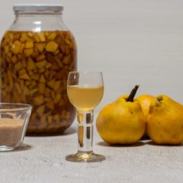 Cuburi de gutui la macert în borcan de sticlă, un bol cu zahăr, un pahar cu lichior și gutui proaspete