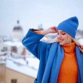 O femeie frumoasă care poartă un pulover orange, un palton albastru și un fes de culoare albastră pentru a ilustra ce imprimeuri și culori se poartă în iarna anului 2023 - 2024