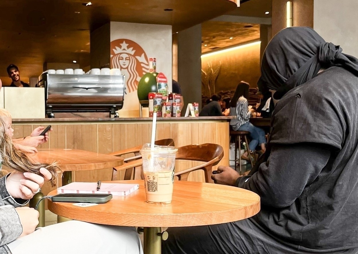 Kanye West, cu o bluză neagră legată pe cap, la o masă, într-o cafenea