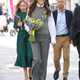 Kate Middleton într-o cămașă albă, o vestă gri și o pereche de pantaloni gri în timp ce are în mână un buchet de flori și se plimbă alături de mai mulți oficiali prin Bracknell