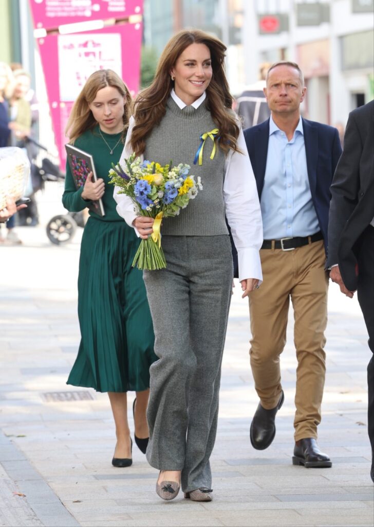 Kate Middleton într-o cămașă albă, o vestă gri și o pereche de pantaloni gri în timp ce are în mână un buchet de flori și se plimbă alături de mai mulți oficiali prin Bracknell
