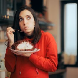 O femeie frumoasă care poartă un pulover roși și mănâncă dintr-o prăjitură cu ciocolată pentru a ilustra ce poți face pentru reducerea poftei de mâncare