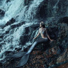 Fată frumoasă cu coadă de sirenă stă pe o stâncă aproape de o apă curgătoare