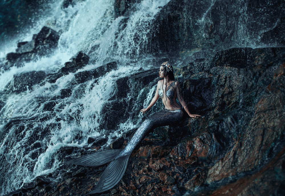 Fată frumoasă cu coadă de sirenă stă pe o stâncă aproape de o apă curgătoare