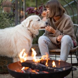 O tânără care stă în curte, pe un scaun, cu un foc aprins, alături de un câine