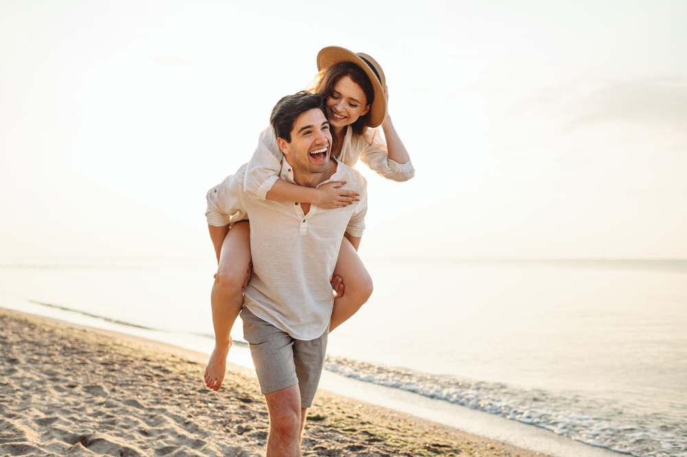 Cuplu care se plimbă pe plajă într-o zi însorită și cei doi zâmbesc
