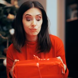 O femeie frumoasă care poartă un pulover roșu și ține în brațe o cutie roșie de cadou în timp ce pare surprinsă