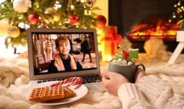 O femeie frumoasă care stă într-un pat și bea ciocolată caldă cu bezele în timp ce se uită la unul dintre cele mai cunoscte filme de Crăciun din toate timpurile