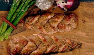 Două bucăți de friptură din mușchiuleț de porc împletit alături de sparanghel verde și ceapă roșie