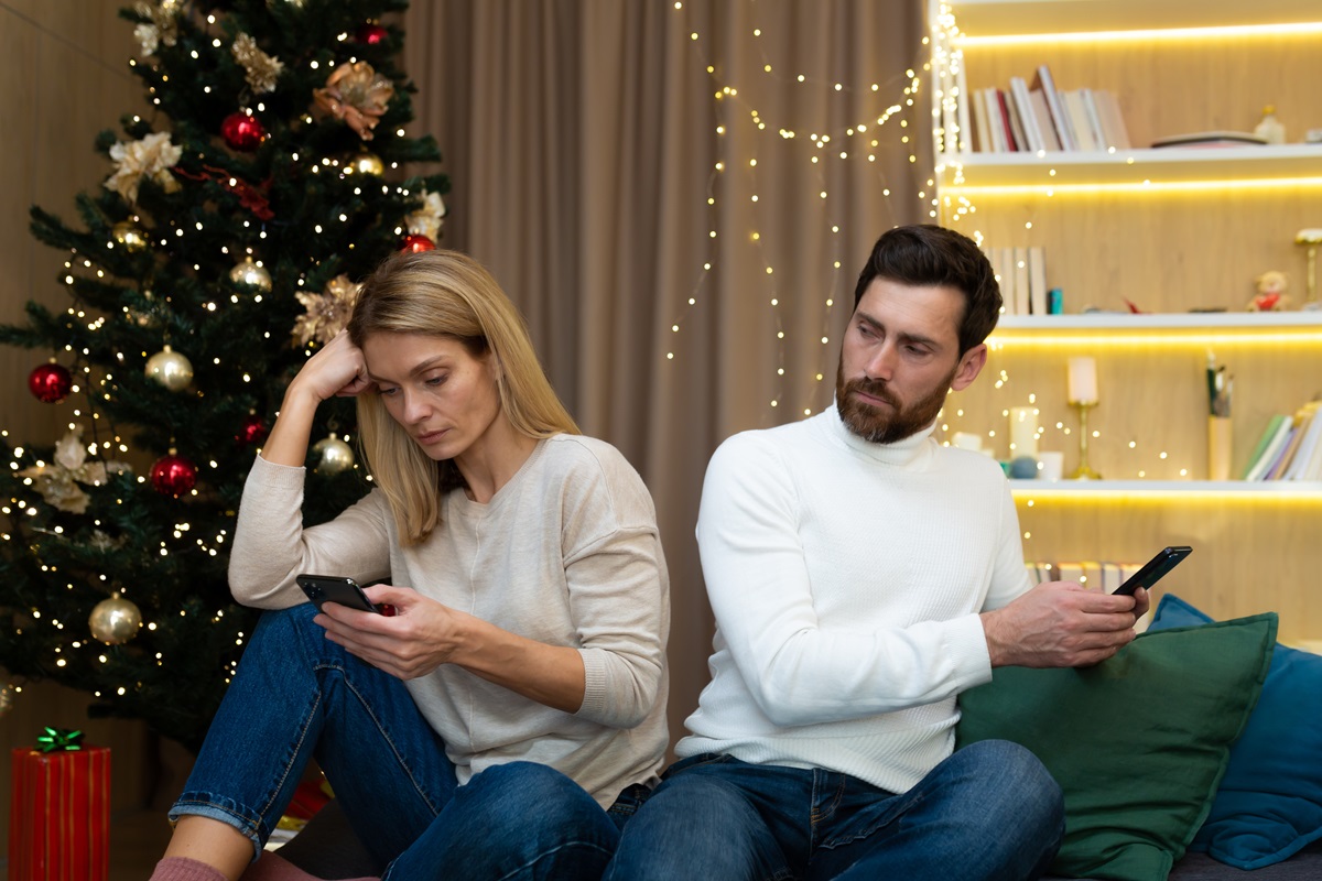 Un cuplu de îndrăgostiți care stau spate în spate în fața unui brad de Crăciun și citesc mesaje de pe telefon
