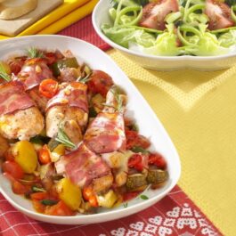Pulpe de pui înfășurate în bacon și așezate peste legume în vas alb, alături de tacâmuri și un bol cu salată