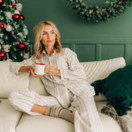 O femeie frumoasă care paortă o pijama albă și stă singură pe o canapea în fața bradului de Crăciun