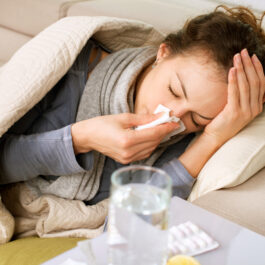 O femeie care este răcită, stă pe canapea și își suflă nasul