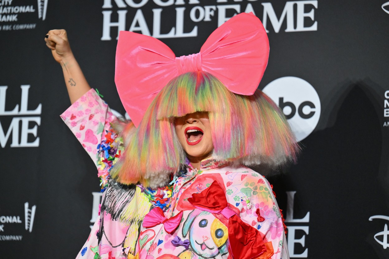 Sia, cu o perucă multicoloră și o fundă mare roz pe cap