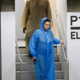 Bianca Censori care coboară scările într-o parcare subterană și poartă o pelerină de ploaie albastră, ea este urmată de Kanye West