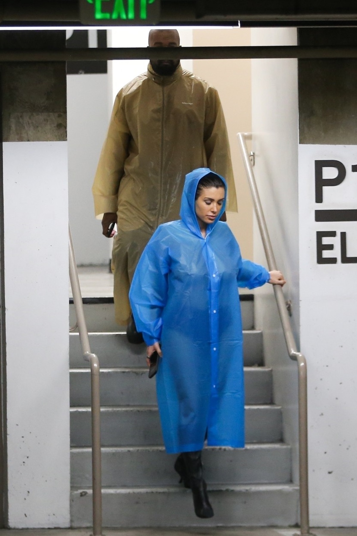 Bianca Censori care coboară scările într-o parcare subterană și poartă o pelerină de ploaie albastră, ea este urmată de Kanye West