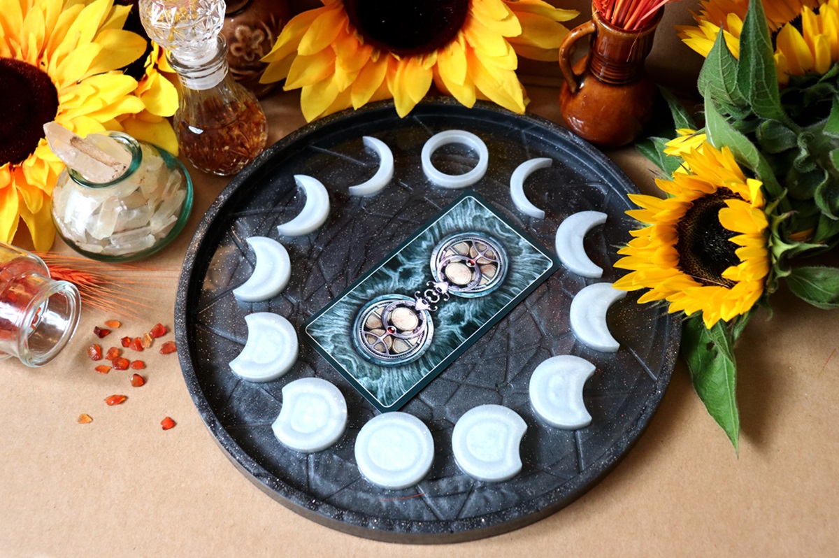 O tavă pe care se află două cărți de tarot și mai multe lumânări albe care ilustrează fazele lunii, înconjurată fiind de mai multe flori de Floarea-Soarelui