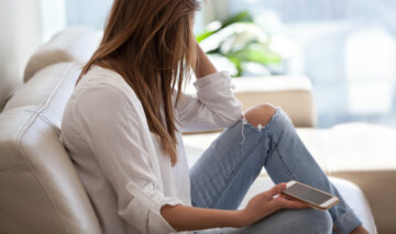 O femeie tristă, care stă singură pe canapea, cu telefonul în mână
