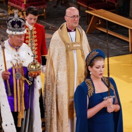 Penny Mordaunt într-o rochie albastră în timp ce ține o sabie la încoronarea Regelui Charles, care se află în spatele acesteia cu globul și sceptrul de aur