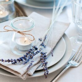 O masă cu tacâmuri frumos aranjate alături de farurfii, o crenguță de lavandă și o luânare decorativă