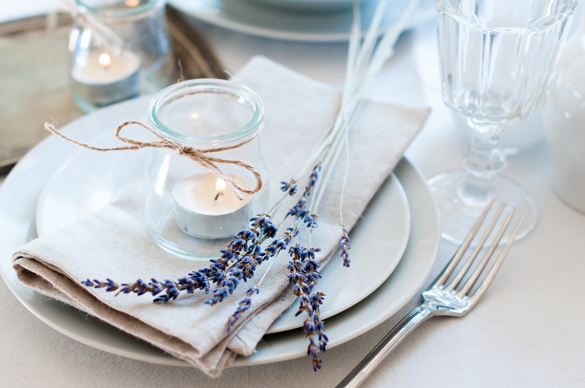 O masă cu tacâmuri frumos aranjate alături de farurfii, o crenguță de lavandă și o luânare decorativă