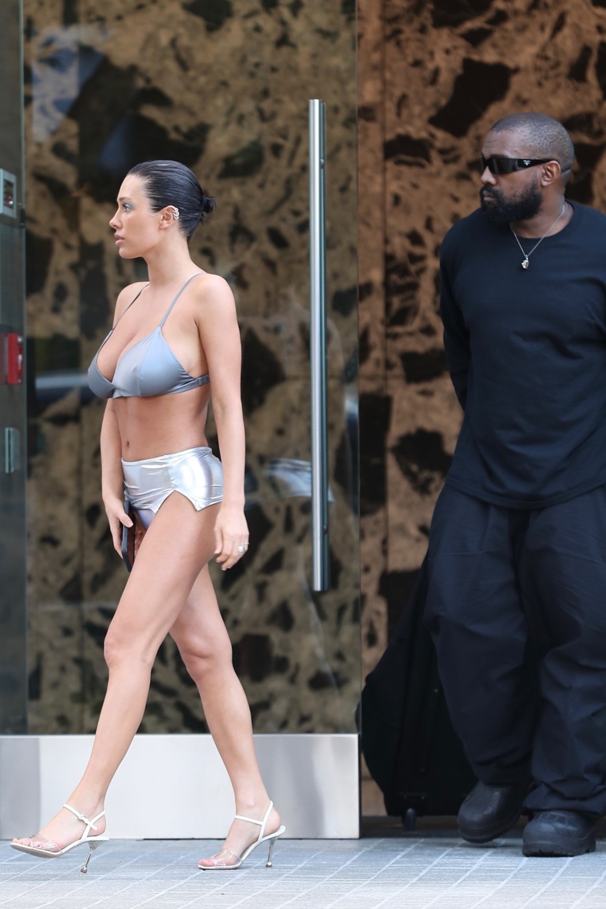 Bianca Censori într-un costum de baie gri în timp ce este urmată de Kanye West la plimbare
