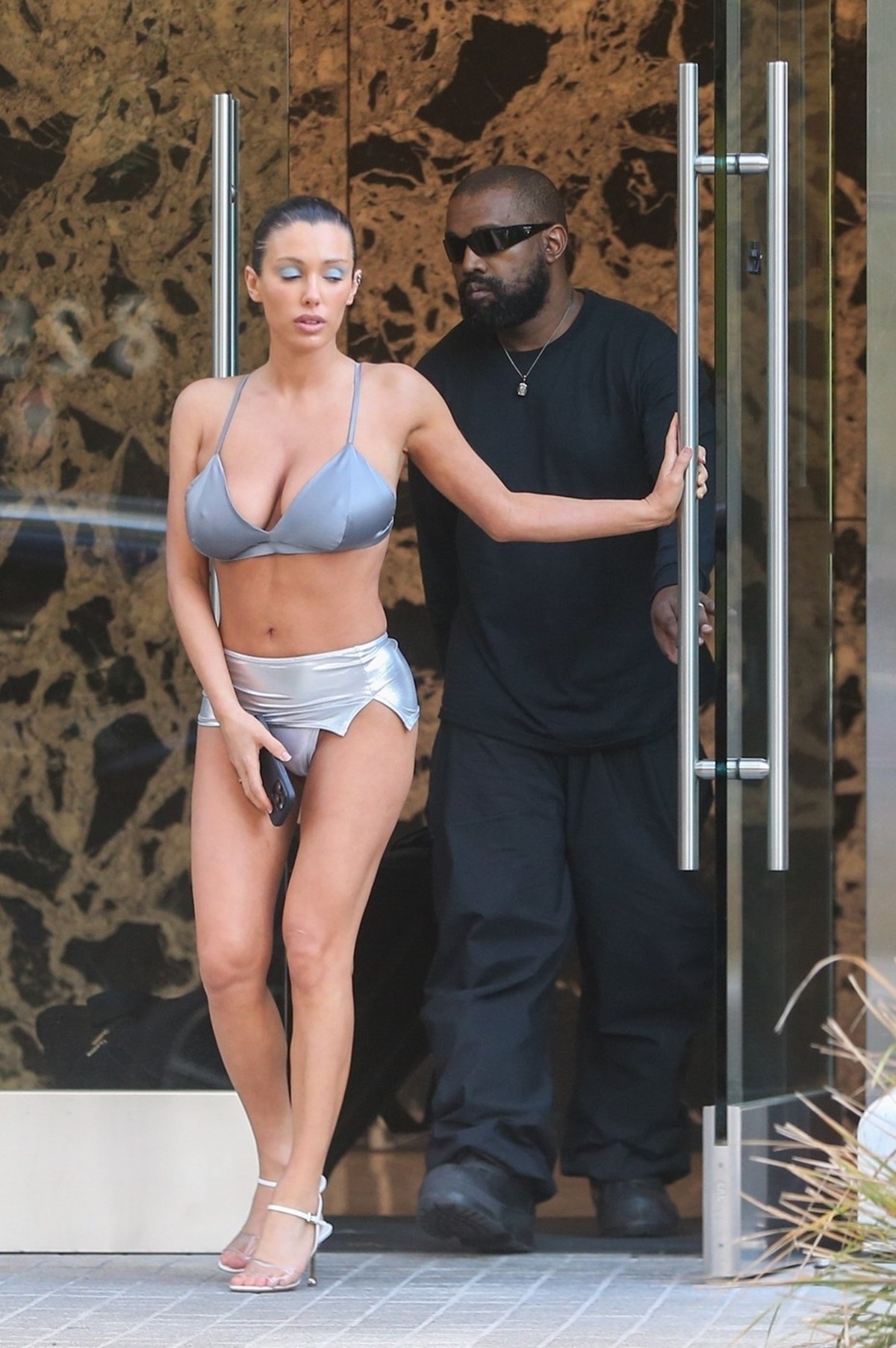 Bianca Censori într-un costum de baie argintiu alături de Kanye West care poartă un costum negru