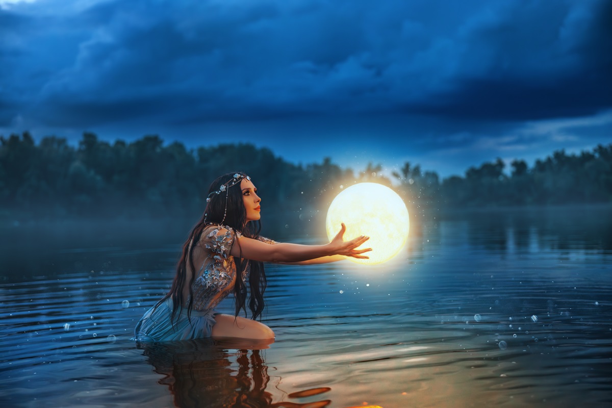 O femeie care poartă o rochie albastră și stă într-un lac în timp ce ține în mână o lună plină pentru a ilsutra fazele lunii în aprilie 2024