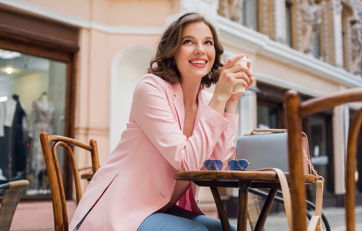 O femeie frumoasă care paortă un sacou roz și stă la o masă pentru a bea cafea